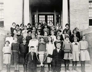 Photograph of group of children on the steps of the school, two of whom are holding a trophy