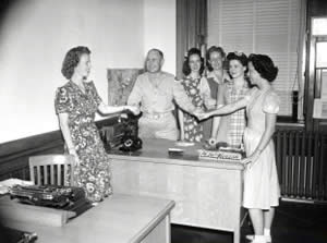 Photograph of military officer behind desk, with five women surrounding him