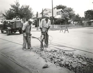 Photograph of two men using a jackhammer to remove the old streetcar rails from the street